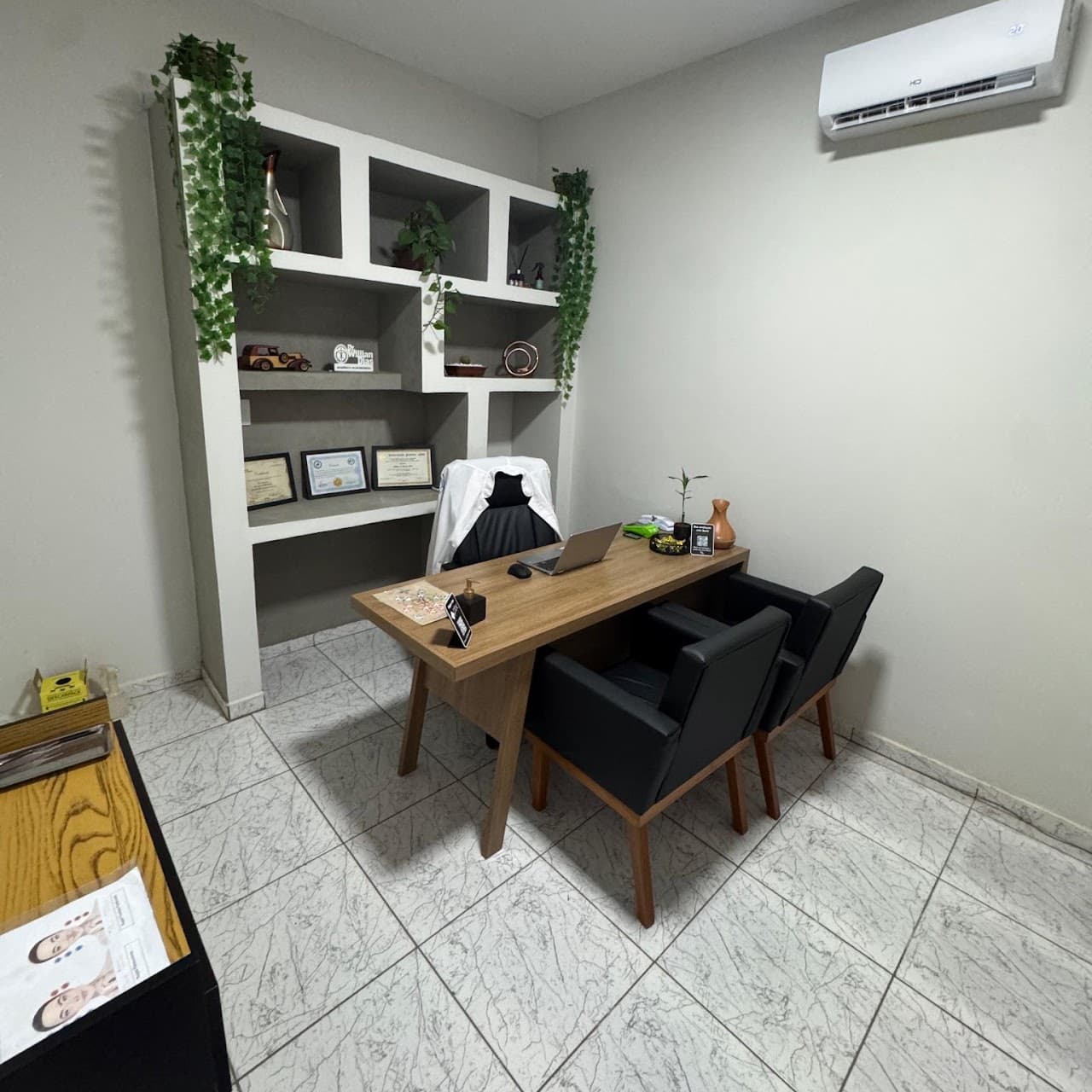 Sala de atendimento - maca em courino preto ao fundo, mesa, poltronas e armário de nichos em alvenaria